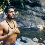 topless man wearing black beaded necklace and blue denim shorts standing on rocky shore during daytime